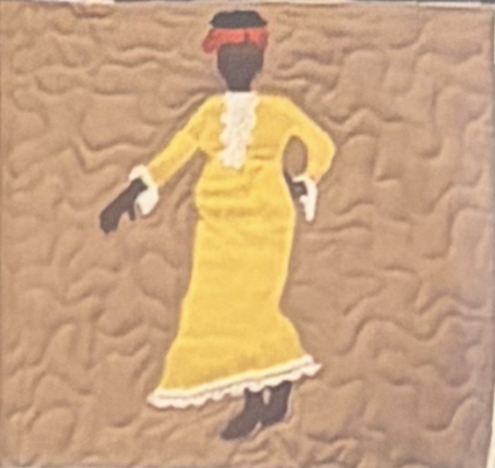 Quilt panel of a Black woman in a long yellow dress with white lace trim and a red headwrap, walking with poise across a warm sand-toned background of hand-stitched ripples