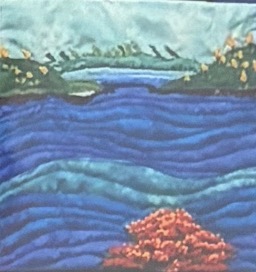 Quilted panel depicting layered blue waves of water with green mangrove shoreline, birds in the distance, and a coral formation in the foreground