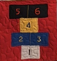 Quilted patch of a hopscotch court rendered in the colors of the Antigua and Barbuda flag: numbered squares 1–6 in white, blue, gold, and black sit on a rich red background with hand-quilted ripples.