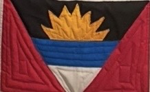 Quilted panel depicting the national flag of Antigua and Barbuda: a golden rising sun over blue and white bands on a black field, flanked by red triangles forming a V shape.