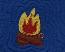 Quilted panel of an appliqued campfire: yellow flames with a red inner flame rise above two brown crossed logs, stitched onto a richly textured deep blue background with concentric quilted ripples.