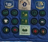 Quilted panel by the 6th Antigua Girl Guide Unit: a deep blue background densely arranged with circular embroidered Girl Guide badges in green, white, and gold, including camp and proficiency badges, framed by a hand-stitched border.