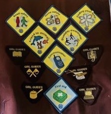 Collection of Girl Guides Brownie badges arranged on a dark surface: yellow diamond-shaped proficiency badges with illustrated symbols (a walking figure, joined hands, magnifying glass, telephone) and brown triangular Brownie patches with crossed axes, a tent, a four-leaf clover, and other emblems, including a 'First Aid' badge.