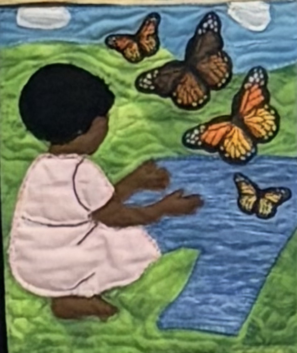 Quilt panel of a young child in a pink dress kneeling beside a pond on green grass, watching four monarch butterflies in flight against a blue sky.