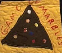 Quilted panel on golden yellow fabric showing a dark triangular playing area scattered with colorful marbles, framed by the words 'GAME OF MARBLES' in red appliqué lettering.