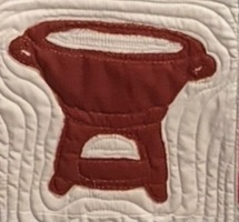 Appliquéd quilt block on cream fabric showing a deep red two-burner camp stove with a rounded pot resting on top, framed by hand-quilted wavy lines.